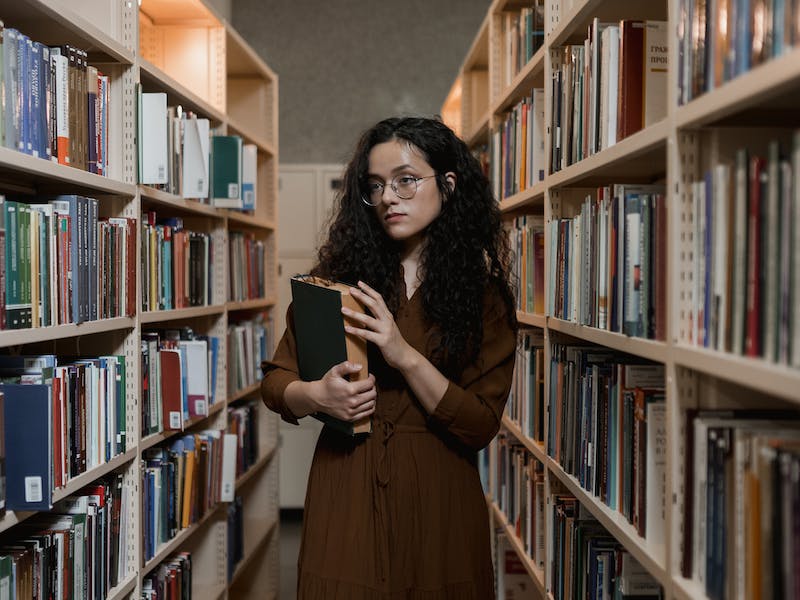 Mujer con libro en la mano caminando entre estantes de biblioteca